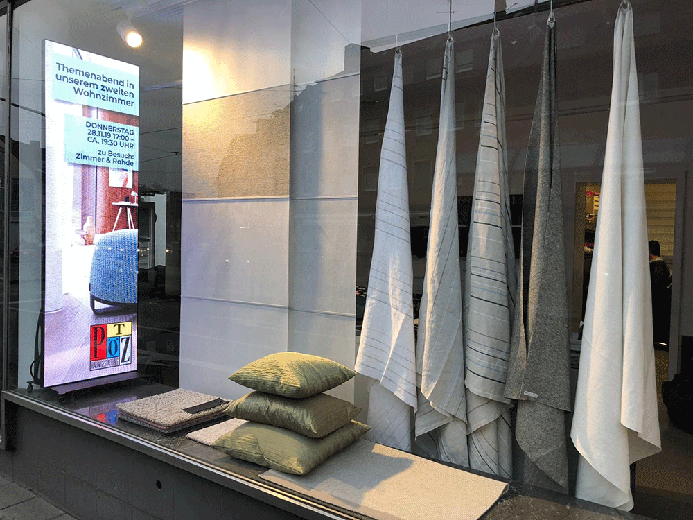 Digitales Roll-Up in einem Schaufenster eines Einrichtungsgeschäfts, neben Stoffmustern und Kissen, wirbt für einen Themenabend.